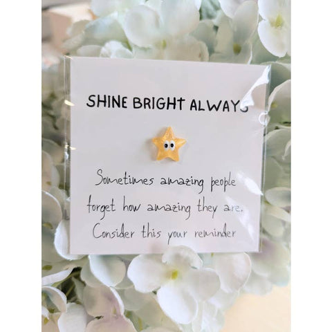 Star-shaped trinket on a card with 'Shine Bright Always' message, surrounded by white flowers.