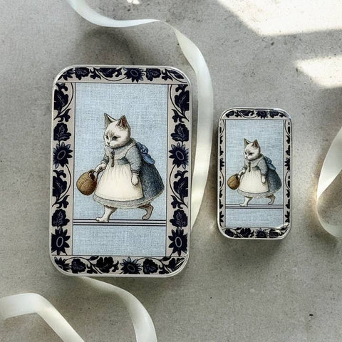 Two tin boxes with illustrations of cats in dresses on a textured surface.