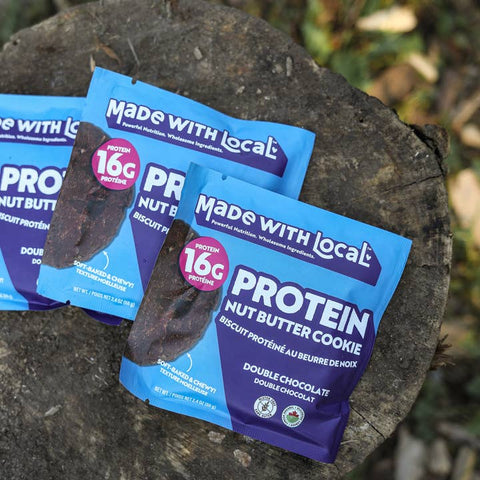 Protein nut butter cookie packages with 'Made with Local' branding on a wooden surface.