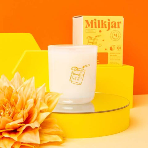 White candle with 'Milkjar' branding on a yellow platform against an orange background