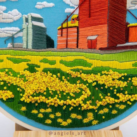 Embroidery of a rural landscape with a red barn, yellow flowers, and blue sky.