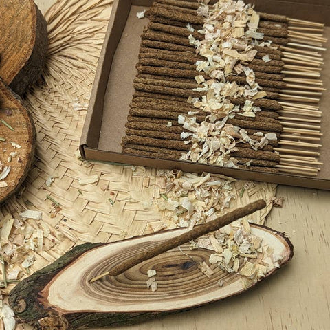 Incense sticks in a box with a wooden dish containing incense ashes on a woven mat.