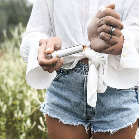 Person wearing a white shirt and denim shorts holding a vape pen with a blurred natural background