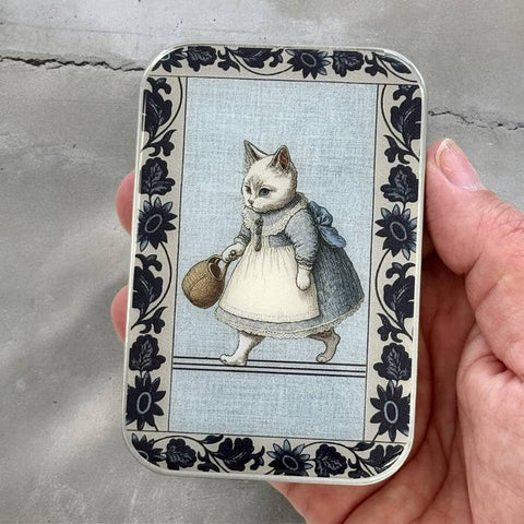 Small tin with a cat illustration holding a basket, held by a hand against a concrete background