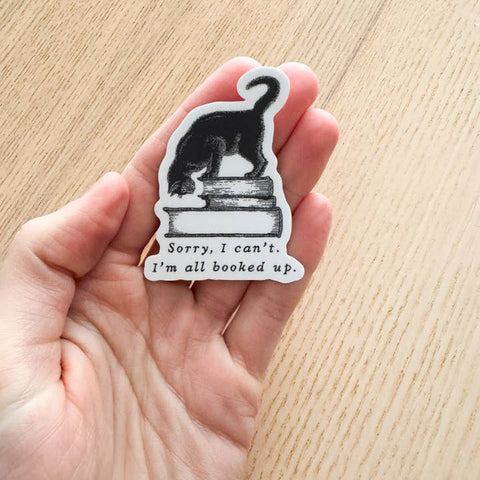 Hand holding a sticker with a cat on books and text 'Sorry, I can't. I'm all booked up.' against a wooden background.