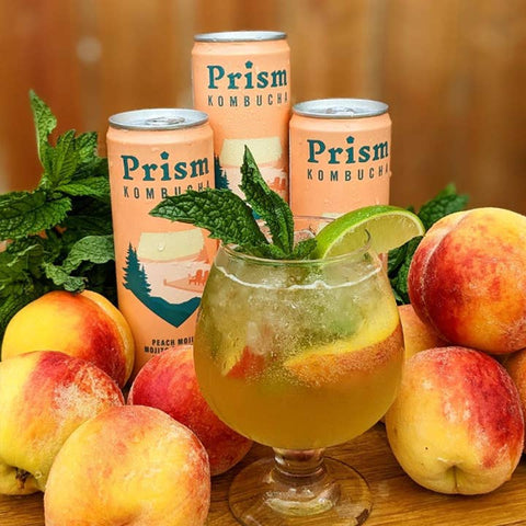 Three cans of Prism Kombucha arranged behind a glass of iced peach-mango kombucha garnished with mint leaves and a lime wedge, surrounded by fresh peaches on a wooden surface with mint leaves in the background.