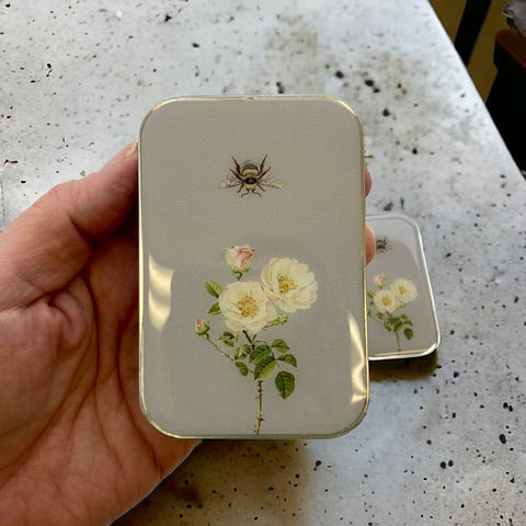Hand holding a small rectangular tin with floral design and bee illustration on a speckled surface.