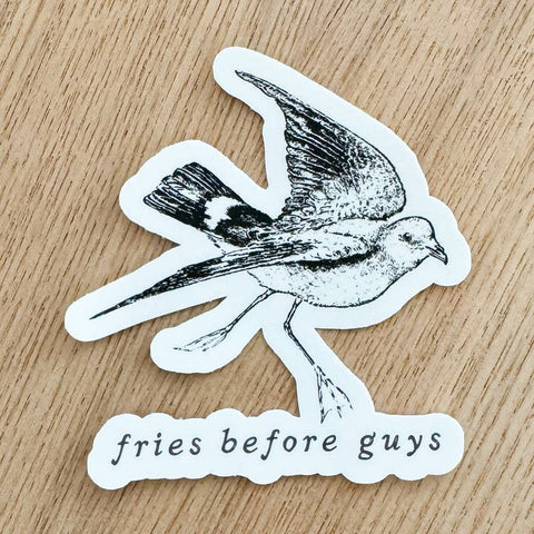 Sticker with a bird design and text 'fries before guys' on a wooden surface