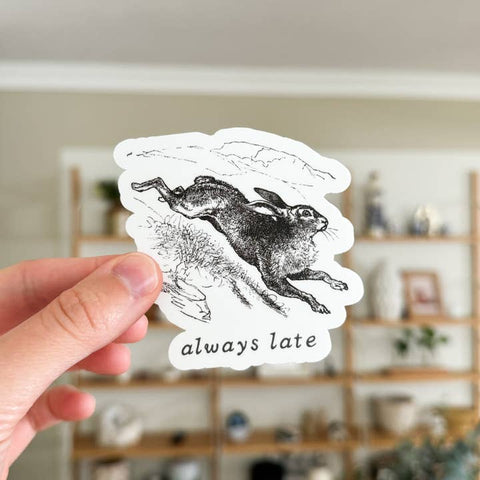 Hand holding a sticker with a black and white illustration of a rabbit and 'always late' text, blurred indoor background