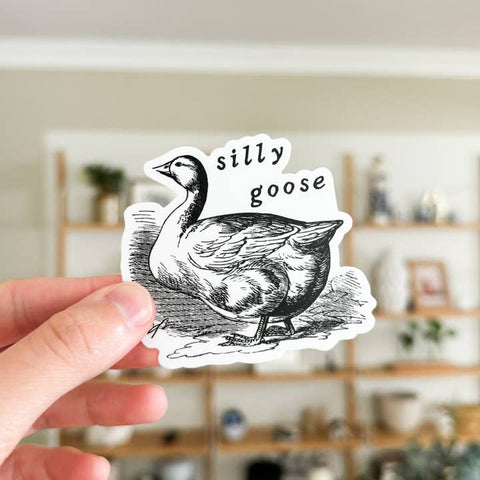 Hand holding a sticker with a goose illustration and 'silly goose' text in a blurred indoor setting.