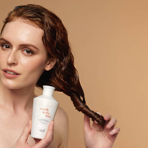 Woman holding a bottle of 'a walk in the sun' shampoo with wet hair against a beige background
