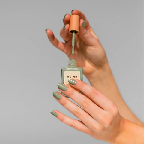 Hand applying green nail polish from a Skind bottle against a gray background