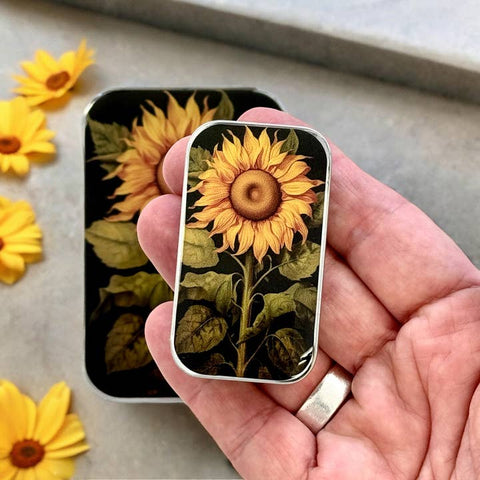 Hand holding a small rectangular tin with sunflower design, surrounded by real sunflowers on a neutral background.