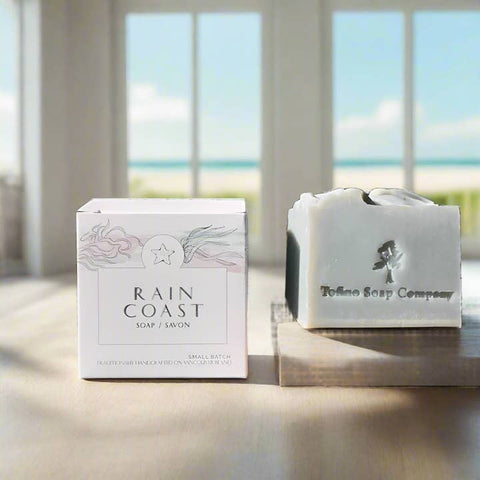Bar of soap and packaging with 'Rain Coast Soap' branding on a reflective surface with a beach view.