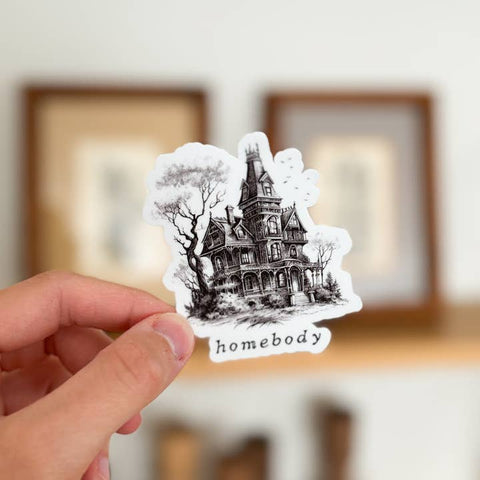 Hand holding a sticker of a haunted house with 'homebody' text against a blurred indoor background.