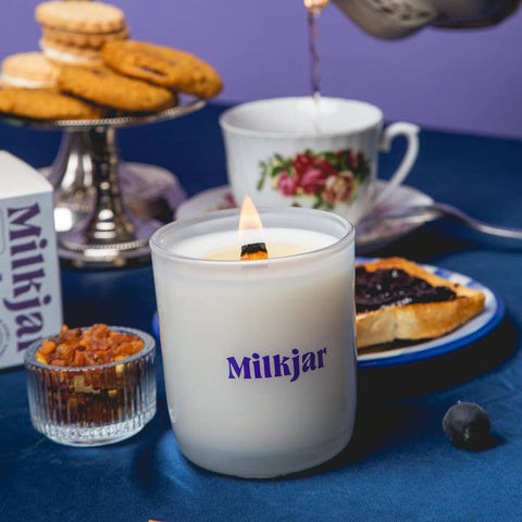 Milkjar candle being lit with a teacup, cookies, and tart in the background on a blue tablecloth.