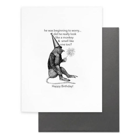 A white greeting card with a black illustrated monkey wearing a party hat and holding a sparkler, with printed text, shown in front of a dark gray envelope on a white background.