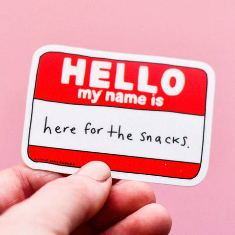 Hand holding a sticker with 'HELLO my name is here for the snacks' text on a pink background