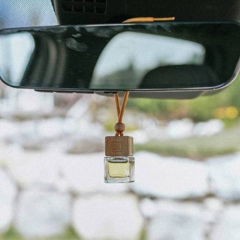 A small glass car air freshener bottle with a wooden cap hangs from a brown cord beneath a rearview mirror inside a vehicle. The background is a blurred outdoor scene with white stones and greenery.