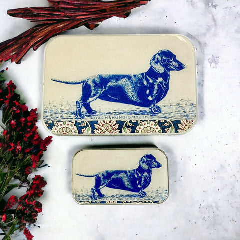 Two tin boxes with blue dachshund illustrations on a textured surface with red branches and flowers.