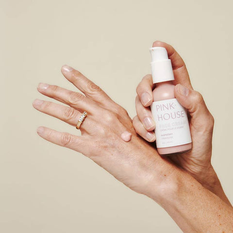 Hand holding a 'Pink House' skincare product with another hand applying it on a beige background