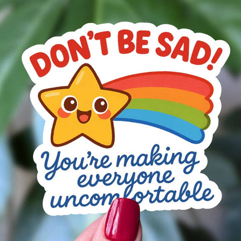 Colorful sticker with a star and rainbow, text 'DON'T BE SAD! You're making everyone uncomfortable', held by a hand with red nail polish.