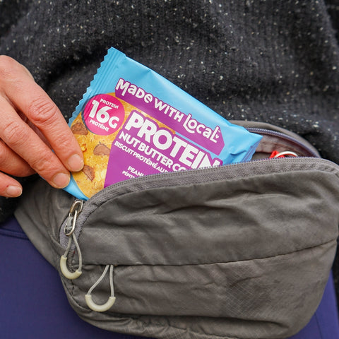 Person holding a protein bar package labeled 'Made with Local' above a gray bag.