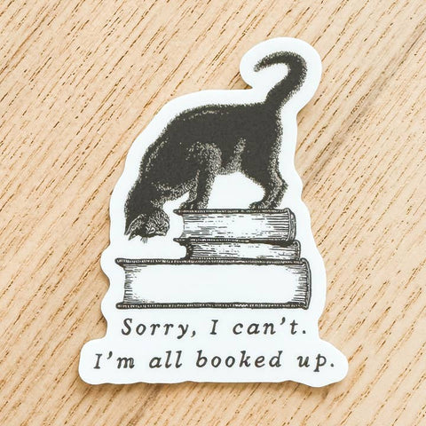 Sticker with a cat on books and text 'Sorry, I can't. I'm all booked up.' on a wooden surface