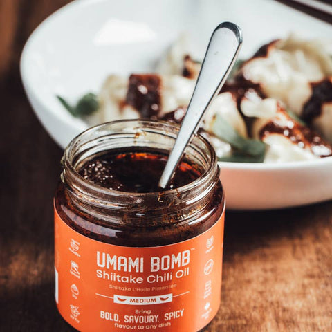 Jar of Umami Bomb Shitake Chili Oil with a spoon in it, placed on a wooden surface with a blurred dish in the background.