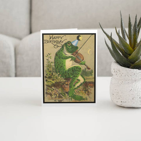 Birthday card with a frog playing violin on a table with a plant in the background
