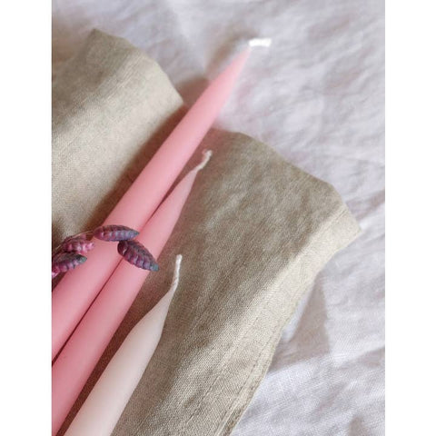 Three taper candles in soft pink, dusty rose, and pale blush tones, arranged on a folded beige linen cloth over a light fabric background.
