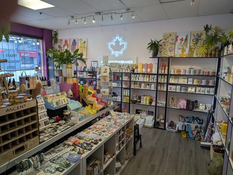 The Local Boom Gift Shop interior with shelves and counters displaying many colorful products, plants, wall art, and a glowing maple leaf sign on the back wall. The store has wood flooring, white walls, and mixed merchandise including packaged goods, books, and accessories.