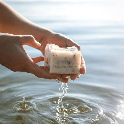 Hand holding a bar of soap over water with a blurred natural background