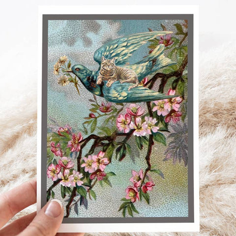 Card with a decorative design of a bird with a cat on its back, surrounded by flowers, held against a soft white background.