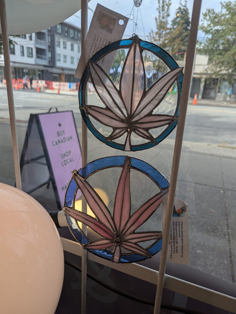 Two circular stained-glass suncatchers with pink cannabis leaves and blue borders hanging on a window, with a blurred street scene in the background.