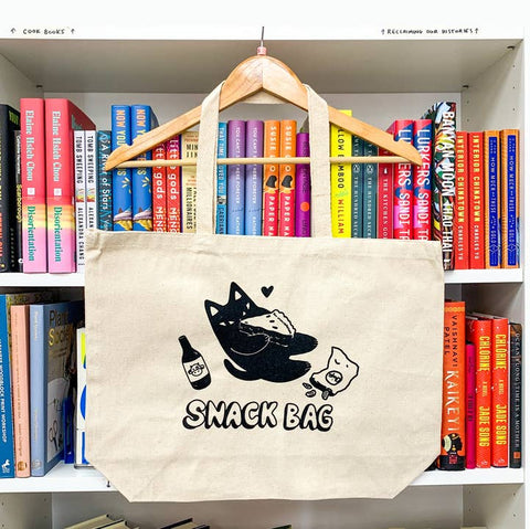 Tote bag with a cat and snack design hanging on a bookshelf filled with books.