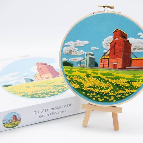 Embroidery hoop with a completed embroidery of grain elevators and a field, next to an embroidered circle and packaging on a white background.