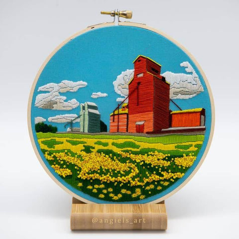 Embroidery hoop with a landscape scene including a red building, green fields, and blue sky.