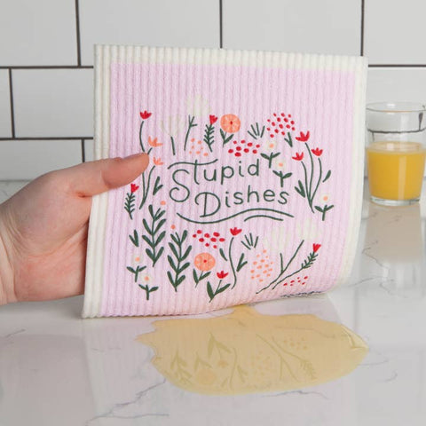 Hand holding a pink dishcloth with floral design and 'Stupid Dishes' text on a kitchen counter.