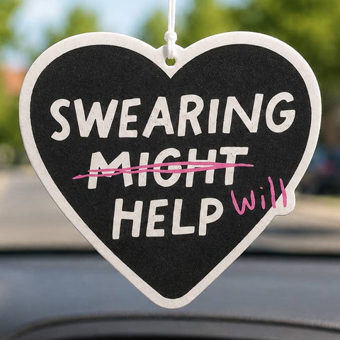 Heart-shaped car air freshener with a black center and white border, hanging in a car window. The text reads 'SWEARING MIGHT HELP' with 'MIGHT' crossed out in pink scribble and 'Will' written in pink script.