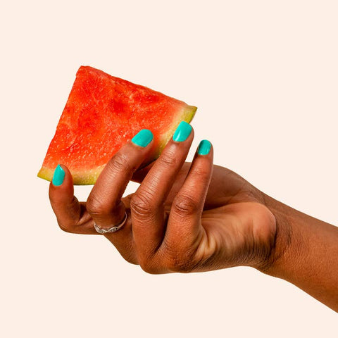 Hand holding a slice of watermelon with turquoise nail polish on a beige background