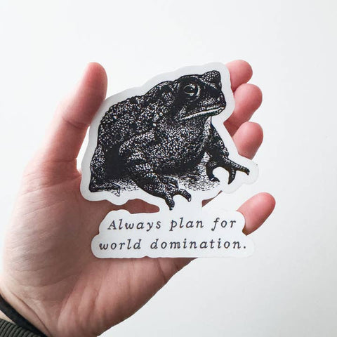 Hand holding a sticker with a frog design and text 'Always plan for world domination' on a white background.