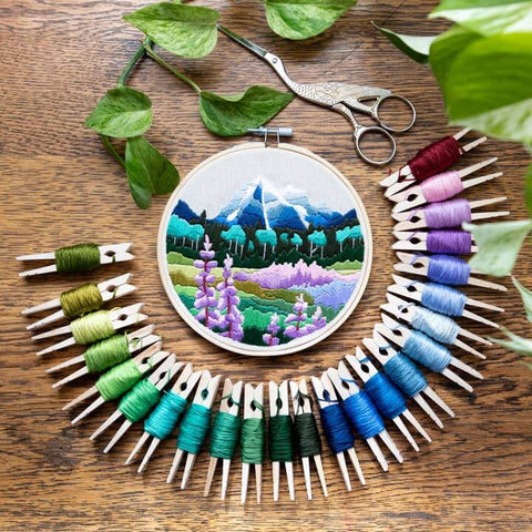 Embroidery hoop with mountain and landscape design surrounded by colorful threads and scissors on a wooden surface.
