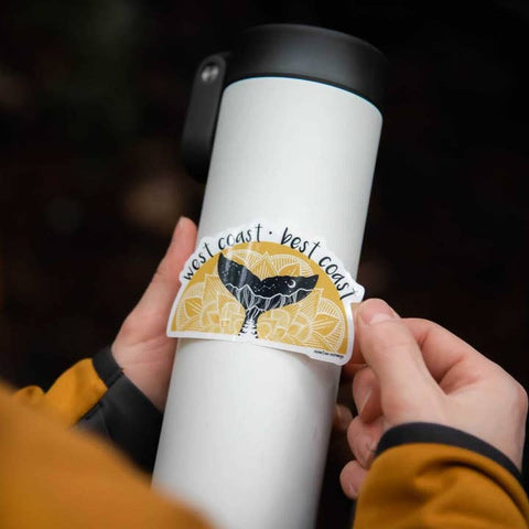 Person holding a white tumbler with a sticker featuring a whale and 'west coast' text.