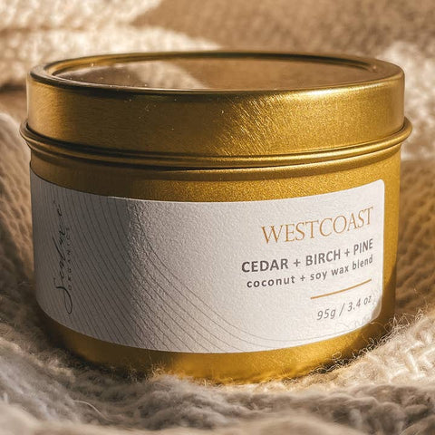 A gold metal candle tin with a white label reading WESTCOAST and Cedar + Birch + Pine, placed on a soft beige textured fabric background.