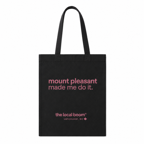 A black canvas tote bag with two shoulder straps, featuring pink printed text that reads 'mount pleasant made me do it.' and 'the local boom vancouver, bc' near the bottom, shown on a plain white background.