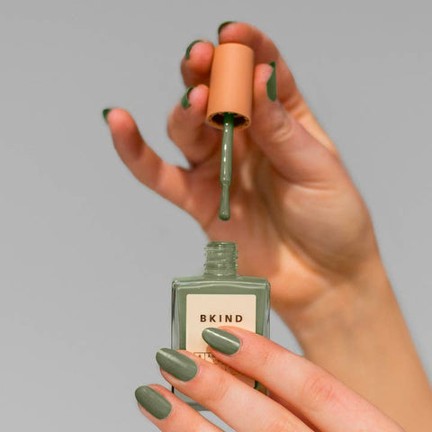 Hand applying green nail polish from a BKIND bottle against a gray background