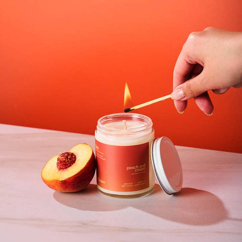 Candle labeled 'Peach and Cream' being lit with a match, next to a peach on a red background.
