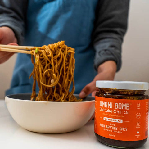Person holding noodles with chopsticks next to a jar of Umami Bomb Shitake Chili Oil.