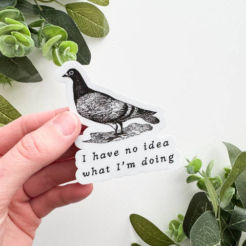 Hand holding a sticker with a pigeon illustration and text 'I have no idea what I'm doing' against a white background with green leaves.